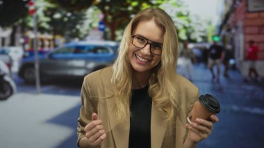 Arka planda arabalar ve binalar olan hareketli bir caddede elinde kahve fincanıyla gülümseyen kadın, kentsel yaşam tarzını ve modasını yansıtıyor..