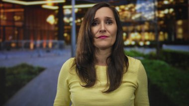 Parlak sarı bir kazak giyen bir kadın şehir merkezinde açık havada enerji dolu bir şekilde jest yapıyor. Bulanık şehir ışıkları mutluluğu ve coşkuyu gösteriyor..