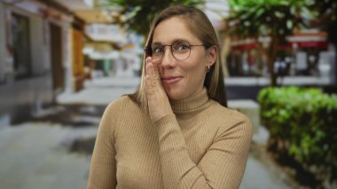 Gün ışığında şehir caddesinde ağaçlar ve binalarla çevrili gözlük ve süveter giyen bir kadın gizli ve sıradan bir atmosfer yayıyor..