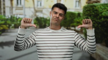 Çizgili tişörtlü İspanyol genç adam güneşli bir şehir caddesinde, şehir ortamında hoşnutsuzluğunu ya da anlaşmazlığını ifade ederek baş parmağını kaldırıyor..