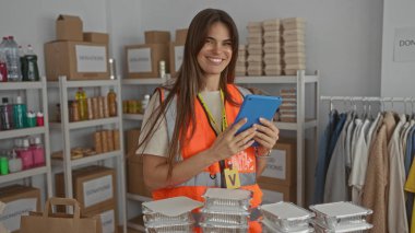 Yansıtıcı yelek giyen genç İspanyol kadın, organize malzeme ve kıyafetlerle çevrili kapalı bir hayır kurumunda tablet kullanıyor..