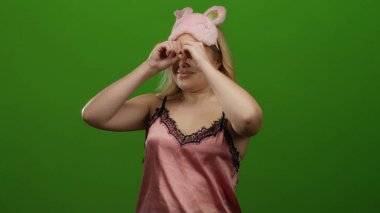 Gece elbiseli kadın elleriyle dürbünlü jest yapıyor, yeşil arka plana pembe uyku maskesi takıyor, neşeli bir ortamda eğlence ve hayal gücünü gösteriyor..