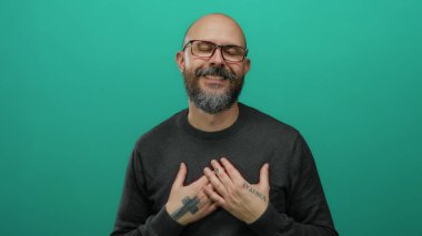 Kel ve sakallı bir adam, yeşil arka plana karşı elleri ve kalbinin üstünde şükranlarını sunuyor, gözlük takıyor ve ellerinde dövmeler gösteriyor..