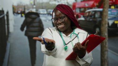 Güler yüzlü kadın doktor, şehir dışındaki açık hava ortamında elinde panoyla elini uzatıyor. Gündüz vakti kameraya dostça bir jest gösteriyor.