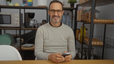 Modern bir iç işyerinde, arka planda rafları olan, gözlüklü ve elinde akıllı bir telefonla güvenle gülümseyen olgun İspanyol bir adam..