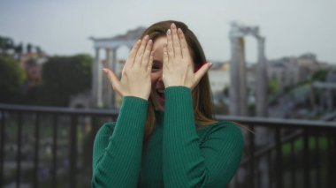 Roma sütunlarına tepeden bakan antik bir binanın yıkıntılarında kadın gülümser ve iki eliyle yüzünü kapatır..