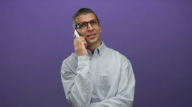 Gözlüklü bir adam akıllı telefonuyla münferit mor arka plan hakkında konuşuyor çizgili gömlek giyiyor, modern iletişim ve gündelik moda tarzı sergiliyor..