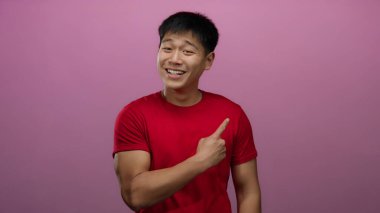 Kırmızı tişörtlü genç Çinli adam başparmağıyla poz veriyor ve pembe arka plan duvarını işaret ediyor, pozitiflik ve onay veriyor..