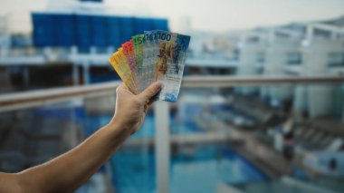 Bir yolcu gemisinde İsviçre frangı banknotları tutan adam, manzaralı bir arka plana karşı bir anlık seyahat ve finansal özgürlük sunuyor..