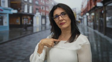 Orta yaşlı İspanyol kadın ıslak şehir caddesinde bulanık vitrinler ve yansıtıcı kaldırımlarla parmak göğse işaret ediyor..