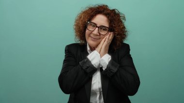Siyah takım elbiseli, kıvırcık esmer saçlı orta yaşlı bir kadın yeşil bir duvar arkasında izole edilmiş bir şekilde uyku jesti yapıyor..