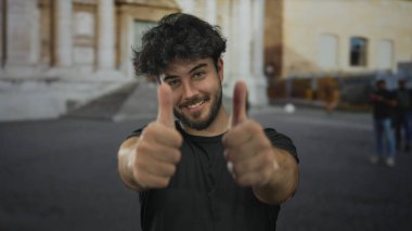 Sakallı genç adam güneşli bir günde arka planda tarihi mimarisi olan şehir ortamında mutluluk ve pozitiflik saçan dışarıda başparmak kaldırıyor..