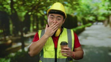 Yansıtıcı yelek ve sert şapka giyen genç bir adam elinde kahve tutarak açık hava parkında şaşırdı..