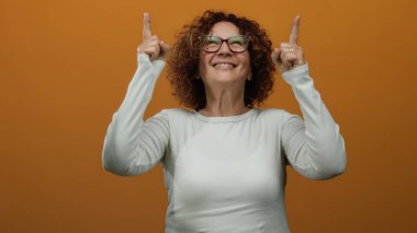Kıvırcık saçlı ve gözlüklü orta yaşlı kadın beyaz gömlek giyiyor, gülümsüyor ve izole edilmiş turuncu arka plana işaret ediyor.