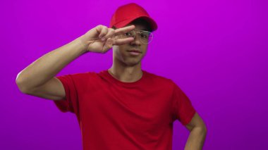 Gözlüklü ve kırmızı gömlekli genç adam mor stüdyo ortamında iki parmağını Ziyaretçi işareti şeklinde tutuyor..