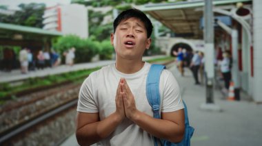 Tren istasyonunda mavi sırt çantalı genç bir adam, kalabalık bir açık hava ortamının ortasında yalvaran bir jest yapıyor..