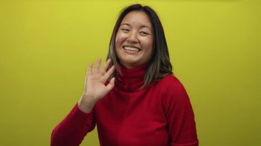 Kırmızı bir kazakla parlak sarı bir arka plana doğru mutlu bir şekilde el sallayan bir kadın izole edilmiş neşeli bir ortamda gençliğini ve pozitifliğini sergiliyor..