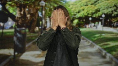 Uzun sarı saçlı, yüzü iki eliyle kaplı bir adam güneş ışığında yeşil park yolu ağaçlarla kaplı; utangaçlık.