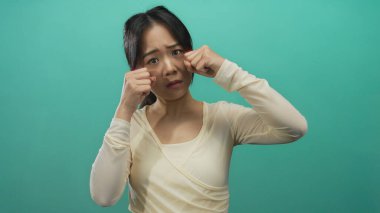 Genç Çinli bir kadın, izole edilmiş yeşil bir arka planda ağlayarak modern, şık bir kıyafet ve düşünceli tavırla stüdyo ortamında duygularını ifade ediyor..