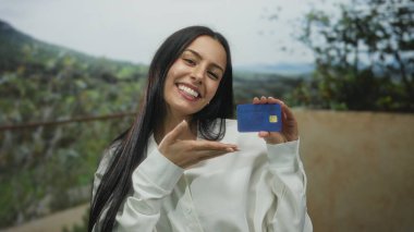 Mavi bir kredi kartını doğal bir ortamda gösteren İspanyol kadın gülümsüyor..