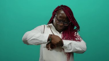 Örgülü ve gözlüklü bir kadın bileğini izole edilmiş yeşil bir duvar arkasına yaslar ve bilek bölgesindeki acıyı ifade eder..