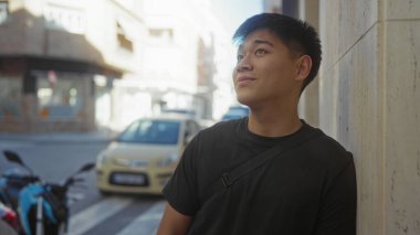 Şehir caddesinde gülümseyen genç Çinli bir adam, arka planda arabaları ve bisikletleri olan bir duvara yaslanmış, kentsel bir ortam öneriyor..