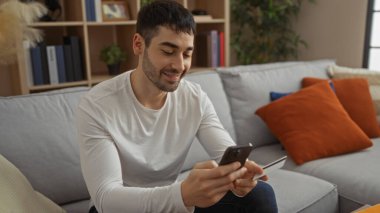 Genç İspanyol bir adam kredi kartını kapalı bir yerde tutuyor, modern oturma odasında gülümsüyor, modern tasarım ve rahat atmosferi olan rahat bir ev ortamını öneriyor..