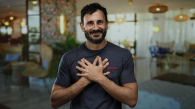 Otel lobisinde elleri göğsünde gülümseyen sakallı İspanyol bir adam, zarif dekorlu bir ev ortamında sıcaklığı ve dostluğu iletiyor..
