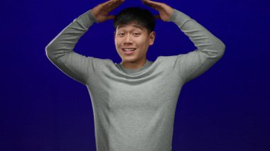 Gri kazaklı genç adam, mavi arka planda elleri başının üstünde şakacı pozlar veriyor..