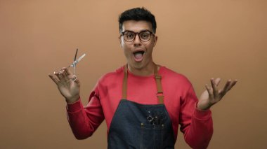 Gözlüklü ve önlüklü genç bir adam kahverengi bir arka plana karşı makas tutuyor ve berberlik mesleğini gösteriyor..