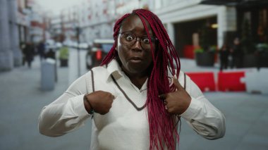 Afro-Amerikalı bir kadın, hareketli bir şehir caddesinde hareketli bir şehir manzarası sergileyerek kendine sürpriz yüz ifadeleri sergiliyor..