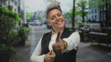 Kısa saçlı, beyaz tişörtlü ve siyah yelekli bir kadın, binaları ve saksıları olan bir şehir caddesinde çifte başparmak işareti yapıyor..