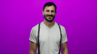 Sakallı, parlak mor arka planda gülümseyen İspanyol bir adam beyaz gömlek ve pantolon askısı giyip renkli ortama karşı neşe saçıyor.