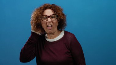 Orta yaşlı, kıvırcık saçlı ve gözlüklü İspanyol kadın. Sanki izole edilmiş mavi bir arka planda dinliyormuş gibi. Merakını ve nişanını dile getiriyor..