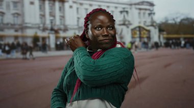 Pembe örgülü bir kadın, tarihi bir bina ve arka planda hareketli bir kalabalıkla güvenli bir şekilde gülümseyerek, canlı bir şehir atmosferini yakalıyor..