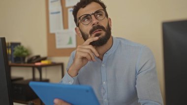 Ofiste sakallı, mavi gömlekli, masasında çalışırken düşünceli ve odaklanmış bir adam..