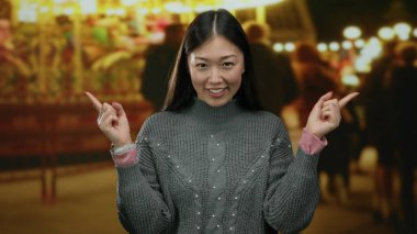 Kadın gülümsüyor ve açık hava fuarına işaret ediyor. Arka planda parlak ışıklar var. Canlı bir akşam atmosferinde..