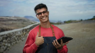 Gözlüklü ve kırmızı kazaklı genç bir adam, elinde tablet, sakin bir caddede gülümsüyor..