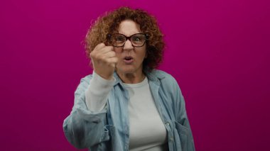Orta yaşlı bir kadın, canlı pembe arka plana karşı öfkesini dışa vuruyor, gözlük ve kot gömlek giyiyor, hayal kırıklığı ve kararlılık duygularını iletiyor.