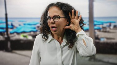 Deniz kenarındaki kıvırcık saçlı İspanyol kadın dikkatle dinliyor, şaşırıyor, plajda gözlük takıyor, merak ve ifadeyi vurguluyor..