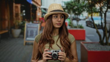 Kameralı genç bir kadın sokakta şapka takıyor. Arka planda ağaçlar ve binalar olan şehir ortamını yakalıyor. Keşif ve macera öneriyor..