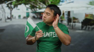 Yeşil gönüllü Asyalı genç adam Pakistan bayrağı taşıyor ve şehir caddesinde dinlerken el kol hareketi yapıyor..