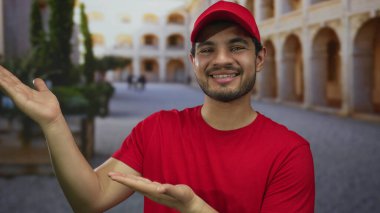 Avlu inşa kemerlerini yumuşak güneş ışığında sunan kırmızı üniformalı Latin asıllı genç adam, dostça bir davet..
