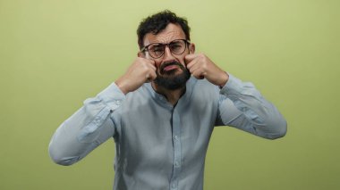 Sakallı ve gözlüklü İspanyol bir adam, sarı arka plandaki üzüntüyü taklit eder. Duygularını yüzüne yakın ifade eder, mavi gömlek giyer..