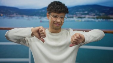 Beyaz kazaklı ve gözlüklü bir adam her iki eli de denize tepeden bakan bir gemide baş parmağını gösteriyor. Onaylamıyor..