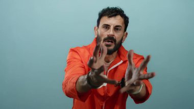 Tutuklu adam turuncu tulum ve kelepçe takıyor, mavi arka planda belirgin bir şekilde jest yapıyor hapsetme ve duygu tasvir eden dramatik bir sahnede.