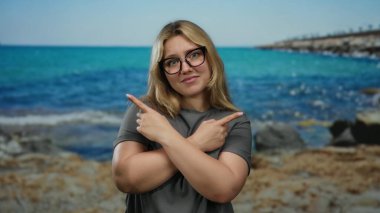 Çapraz kollarla poz veren kadın deniz kenarında farklı yönlere bakıyor. Arka planda çarpıcı mavi deniz ve doğal manzara var..