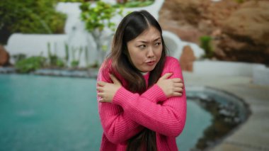 Pembe kazaklı kadın havuz kenarında poz veriyor. Soğuk, gözleri kapalı ve kolları çaprazlanmış bir şekilde sakin bir otelde huzurlu Asyalı güzelliğini sergiliyor.