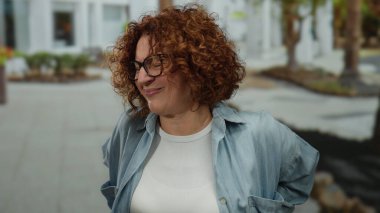 Şehir caddesinde acı içinde kıvırcık saçlı ve gözlüklü orta yaşlı, rahatsız edici İspanyol bir kadın, günlük mavi beyaz giyinmiş, açık şehir manzarası..