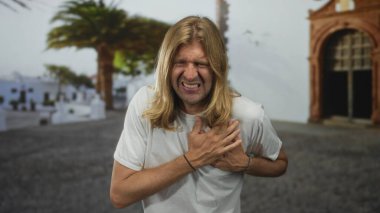 Issız bir sokakta göğsünü sıkarken uzun saçlı, somurtkan ve şaşı sarışın bir adam. Kalp krizi acısı..